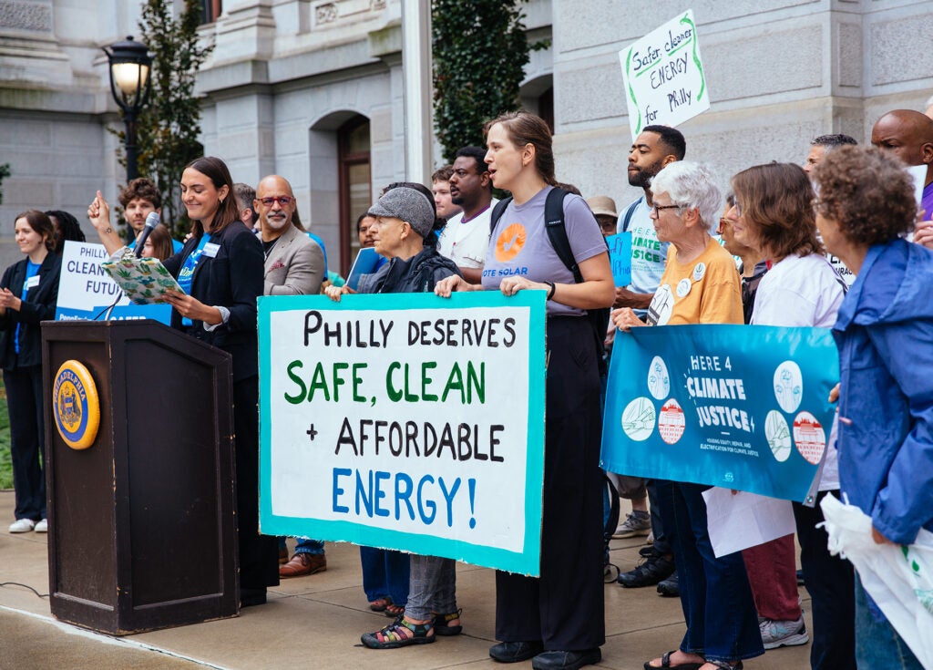 A group of people stand behind a speaker at a podium. Some hold signs including a large one that says "Philly deserves safe, clean + affordable energy!" and "Here 4 climate justice"