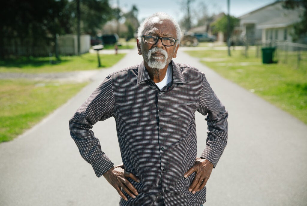 An older man in a checkered shirt with his hands on his hips and a serious expression looks directly into the camera. He is standing in the middle of a residential street.
