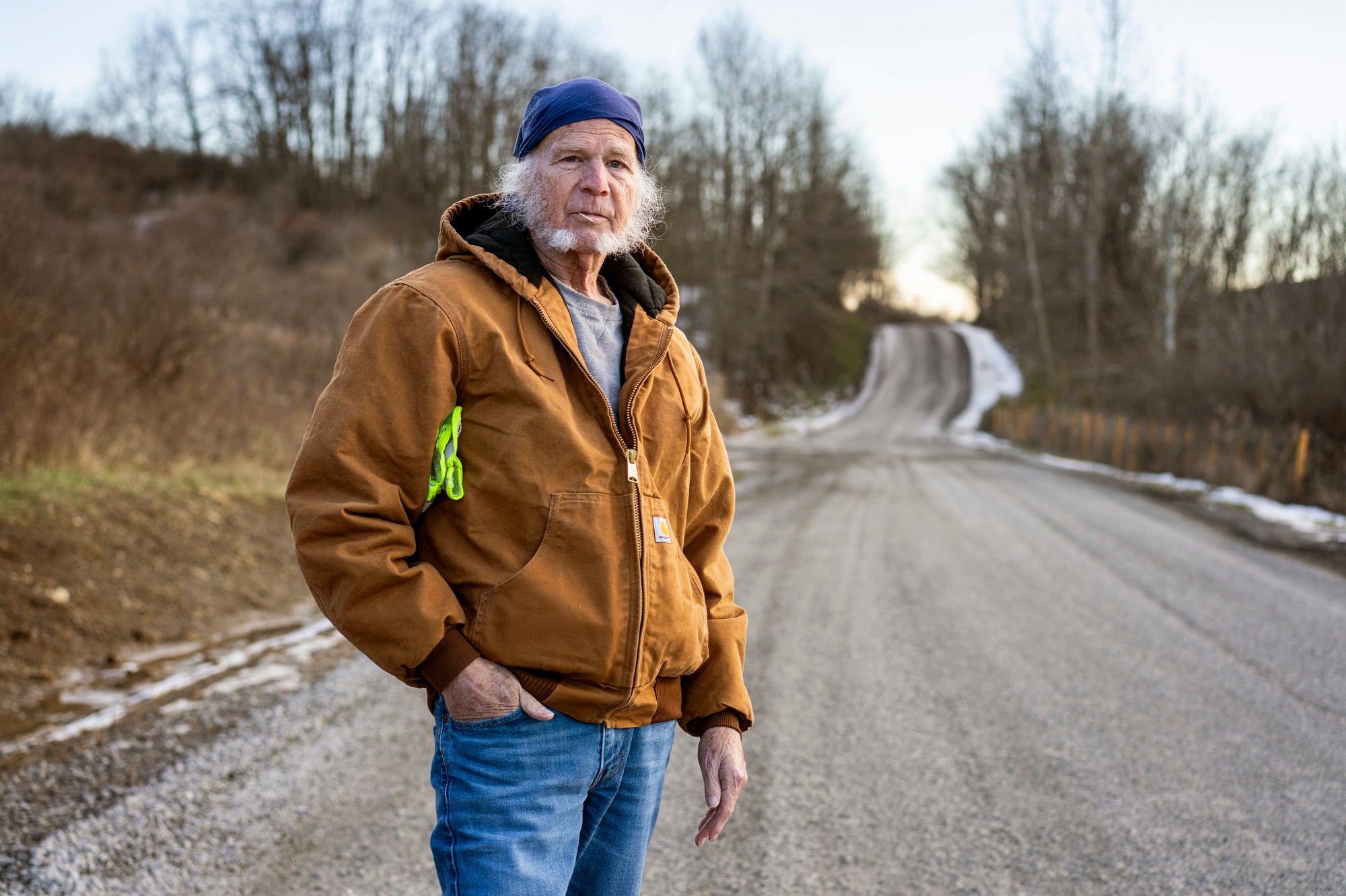 An older man with a blue bandana and tan work coat stands on a rural road in the winter.