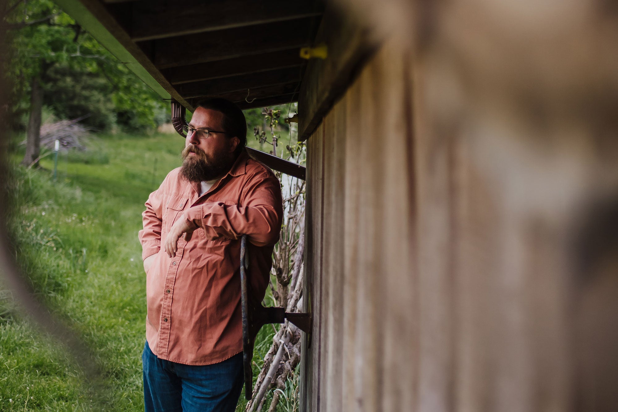 Alex Cole, wearing a peach-colored buttoned-down shirt and blue jeans, leans against the side of a wooden building set in a green, verdant landscape.