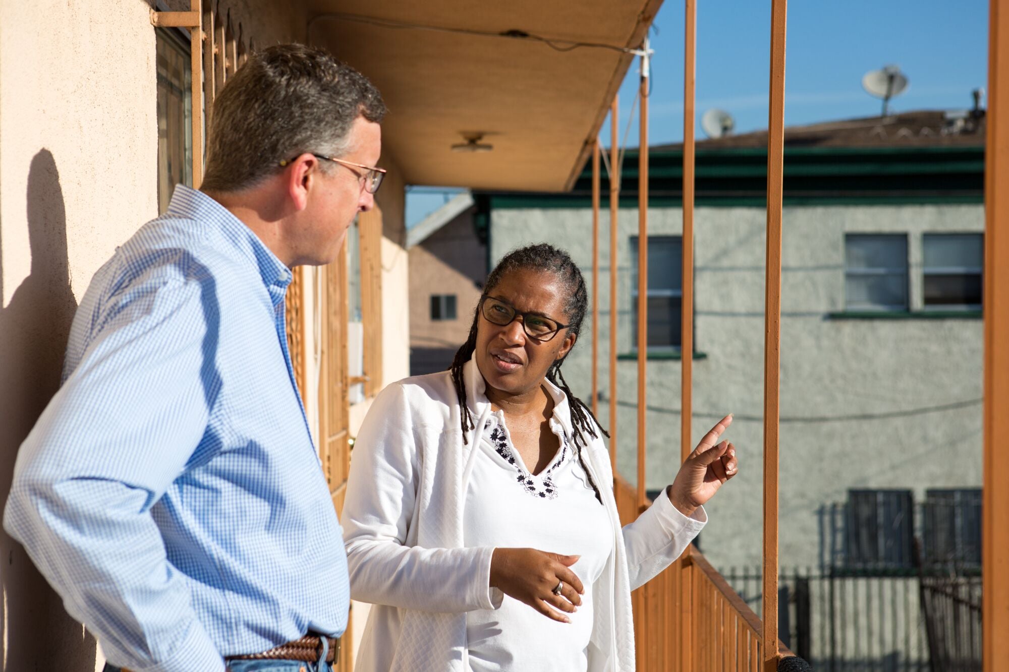 Angela Johnson Meszaros, wearing a white blouse and jacket, speaks to Redeemer Community Partnership President Richard Parks, who is wearing a light blue dress shirt and standing, listening with his hands on his hips. The two are on an exterior, elevated balcony on a clear, sunny day. Meszaros points to the right as she speaks.