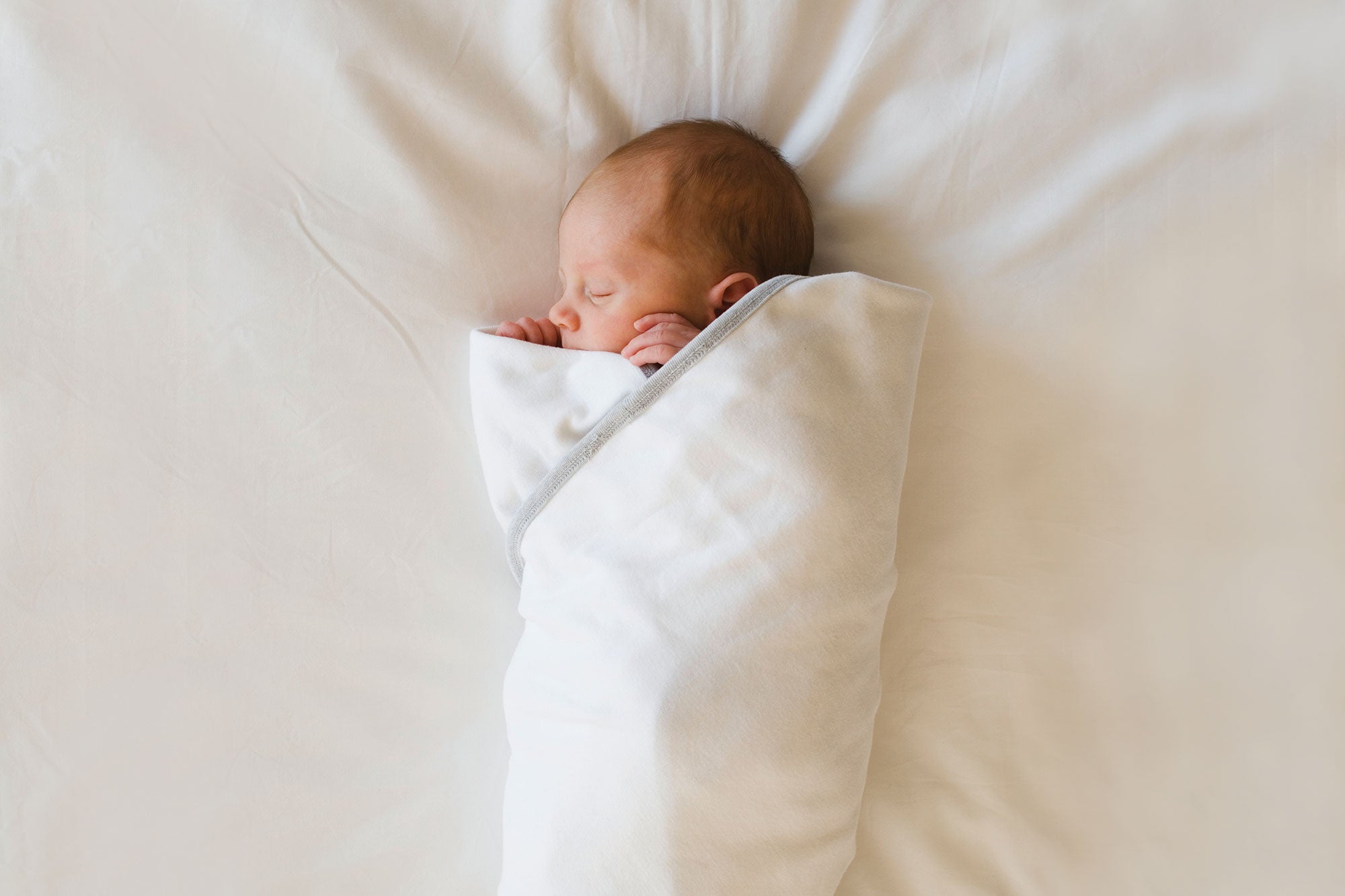 A small baby swaddled in a cream-colored blanket lays peacefully on a bed with cream-colored sheets.