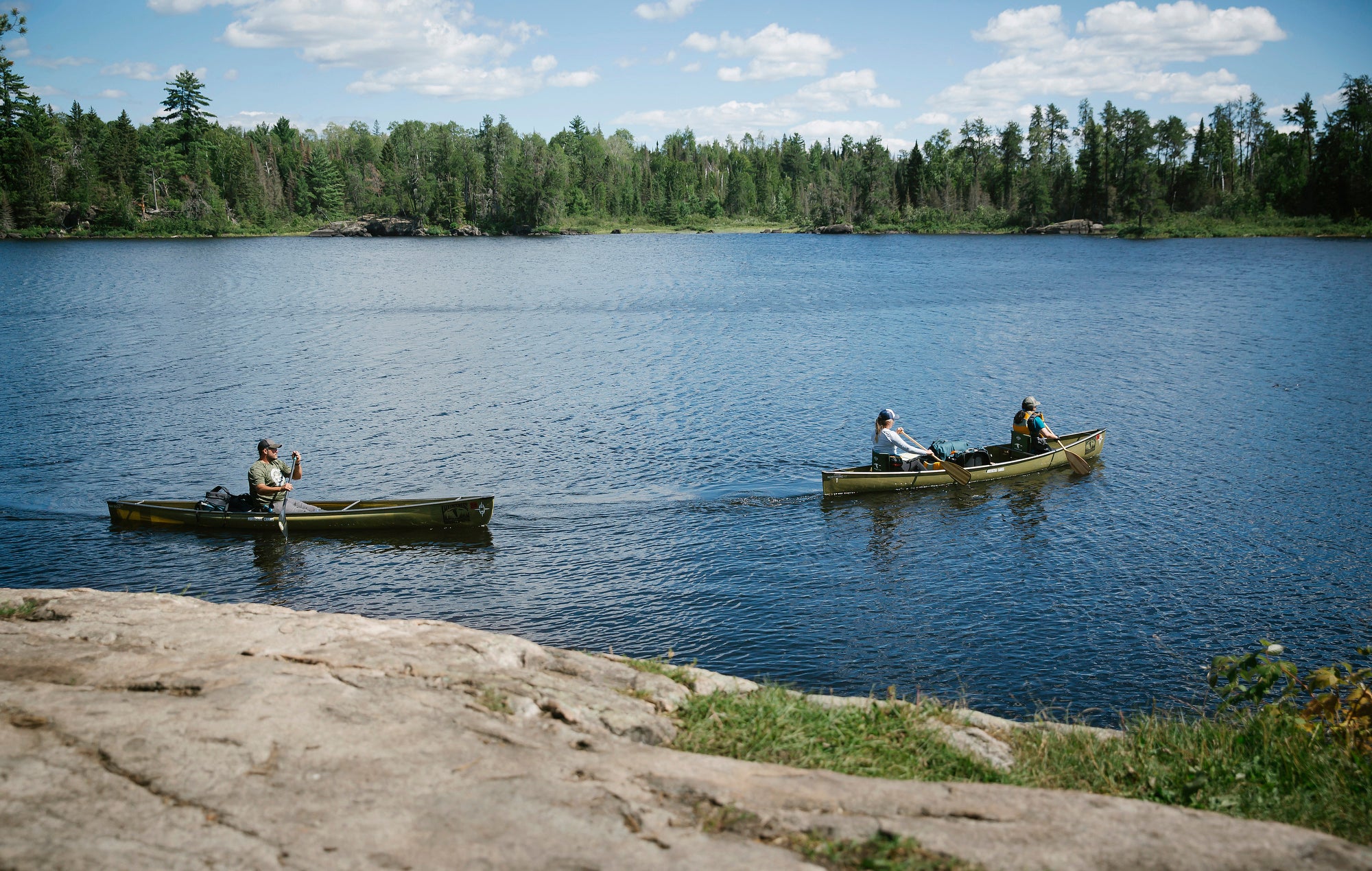 Two canoes, one with two people and one with one, paddle along a lake with a rocky shore in the foreground and a tree lined bank in the background.