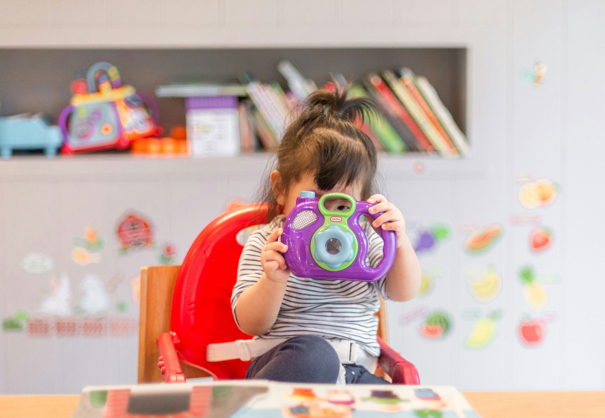 A toddler in a bright red booster seat sits at a table in a playroom, looking closely at a plastic purple camera toy.