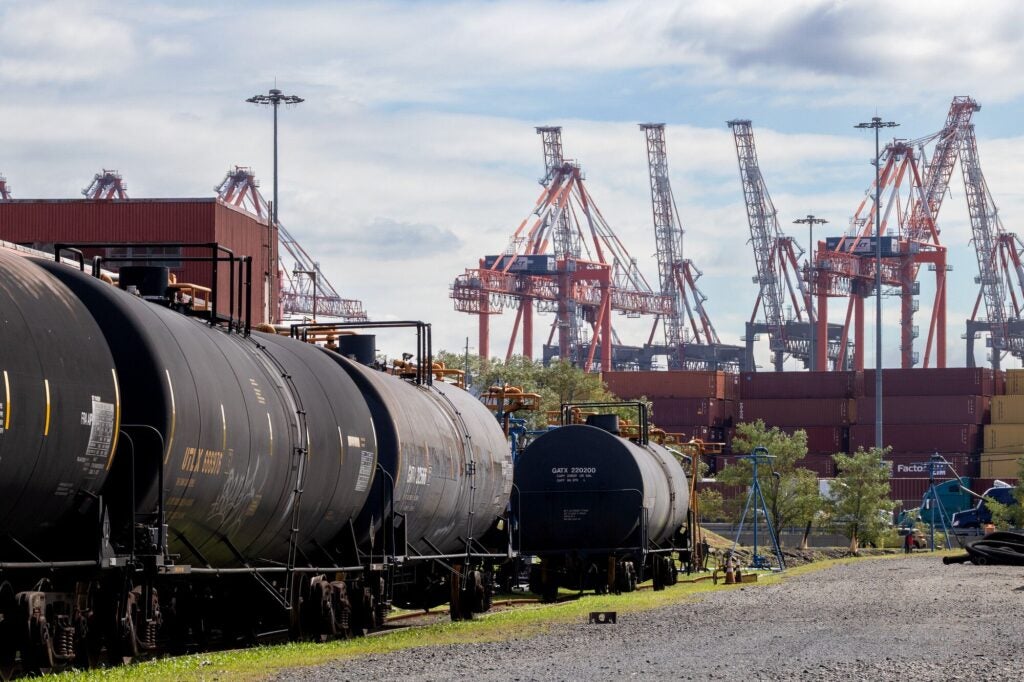 Black, cylindrical train cars on tracks next to stacks of containers and large cranes in the distance.