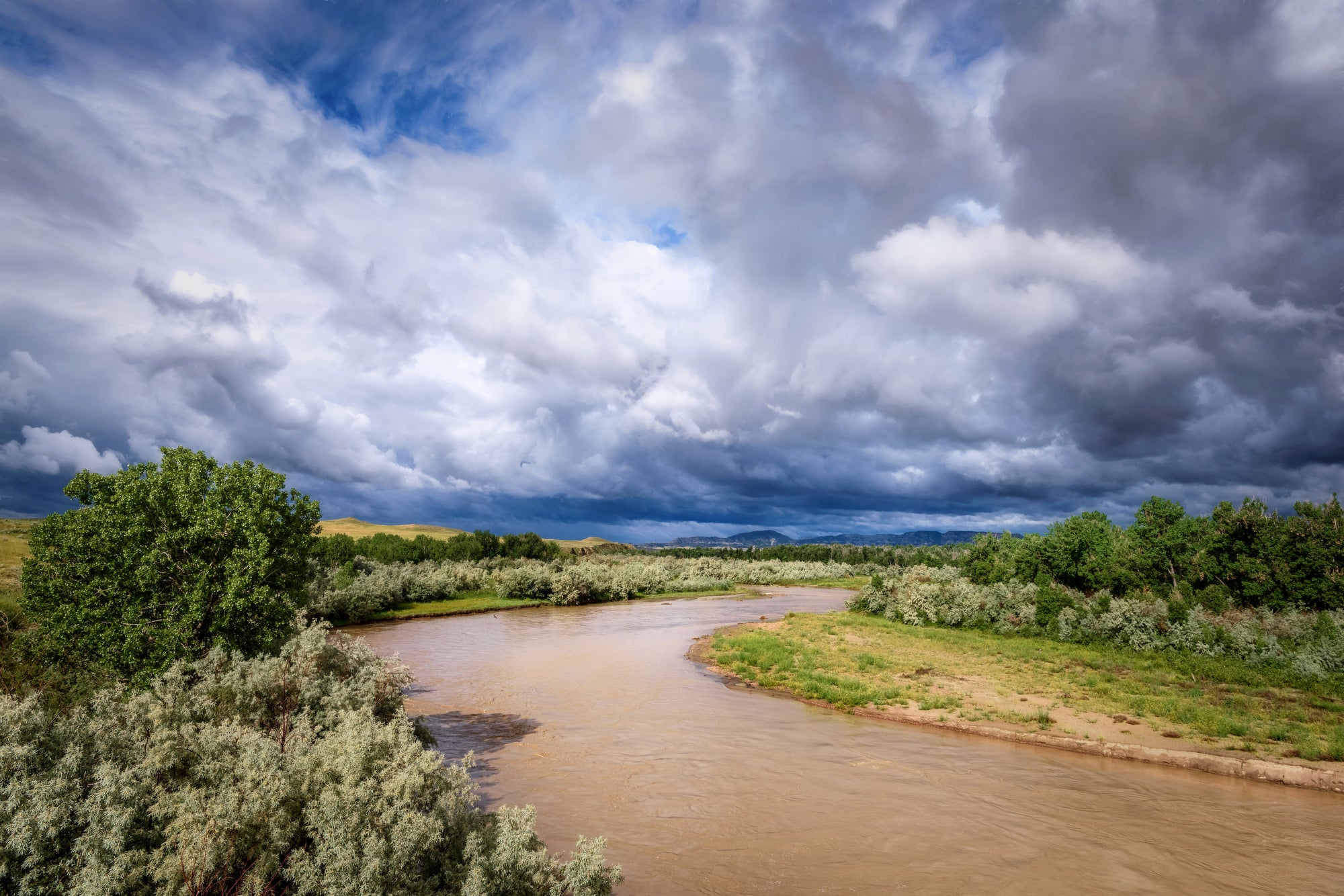 A muddy river snakes through brush and low, grassy hills with large clouds above it.