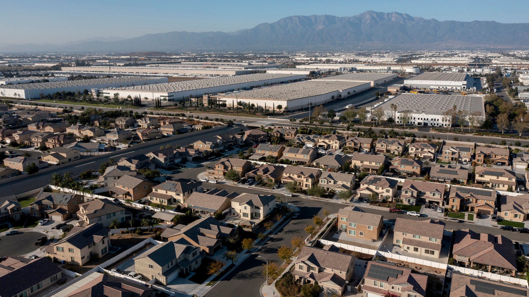Suburban houses in the foreground are next to an expanse of large, white warehouses that stretch into the distance. The horizon is hazy with mountains in the distance.
