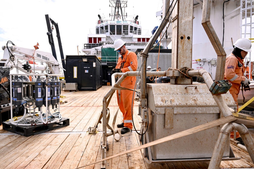 Two men in orange work suits stand on the deck of a ship with pieces of large equipment, including one that looks like a scoop.