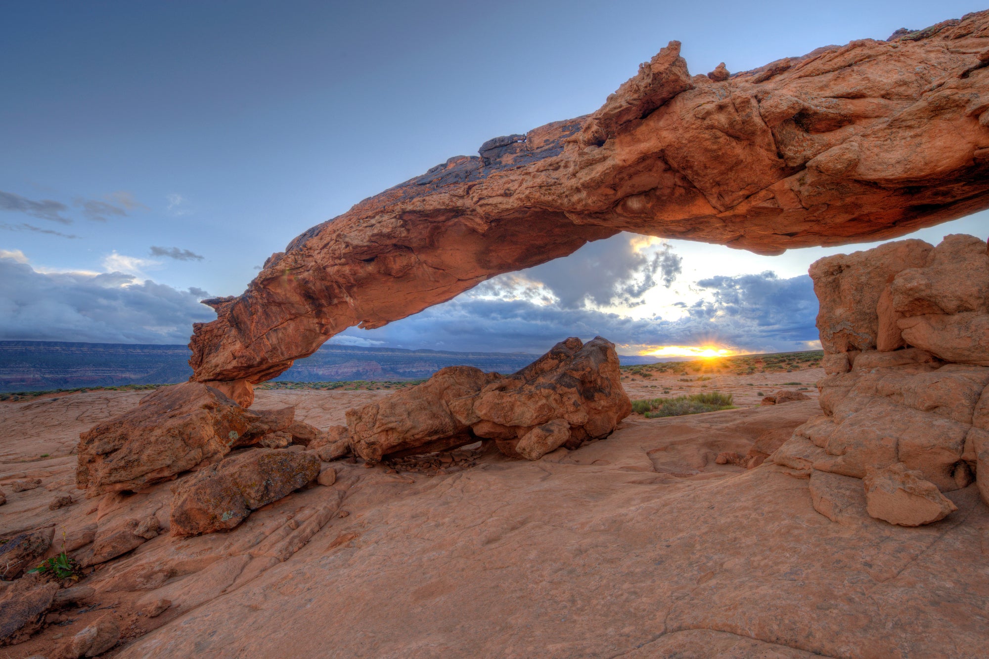A rock arch in a red rock formation overlooking a southwest desert scene with the sun low on the horizon.