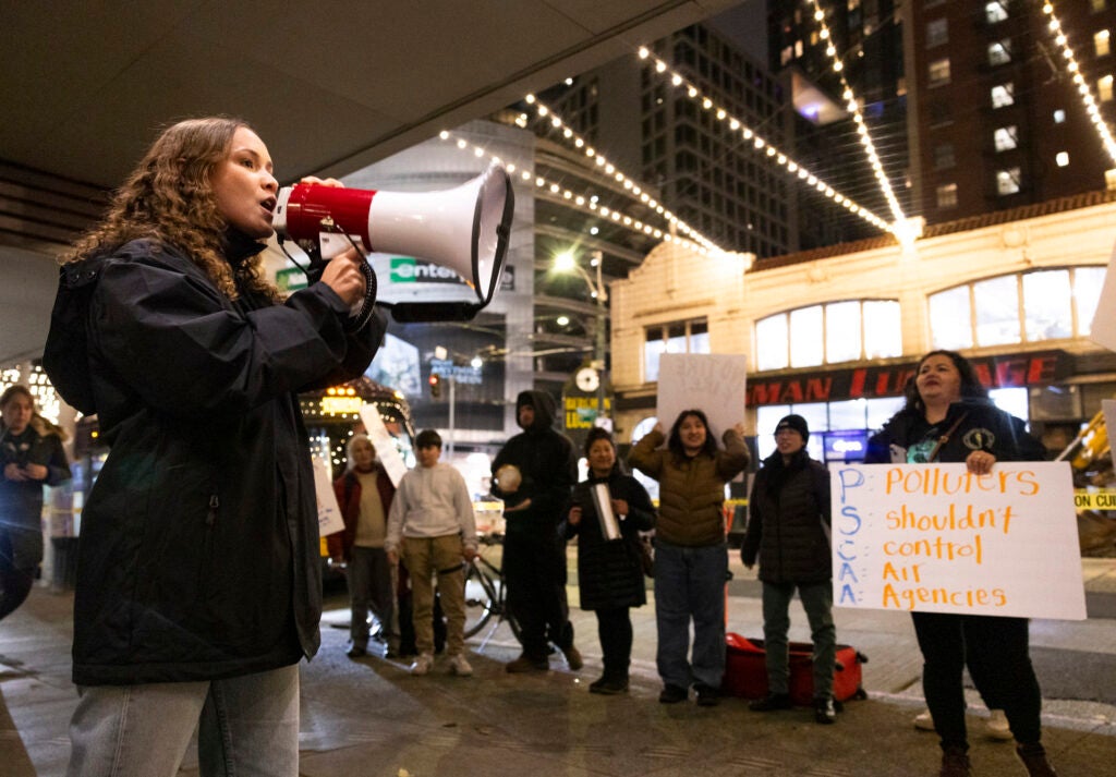 A woman holding a megaphone speaks to a group gathered on a city sidewalk at night. One person is holding a sign that reads: "Polluters shouldn't control air agencies"