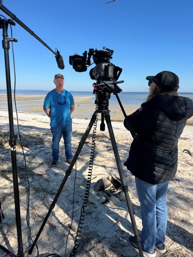 A man in a blue t-shirt, jeans, and a dark-colored cap, stands on a beach on a sunny day, with his hands behind his back, talking into a video camera and a boom microphone, as a woman wearing a black puffy jacket stands behind the video camera watching him speak.