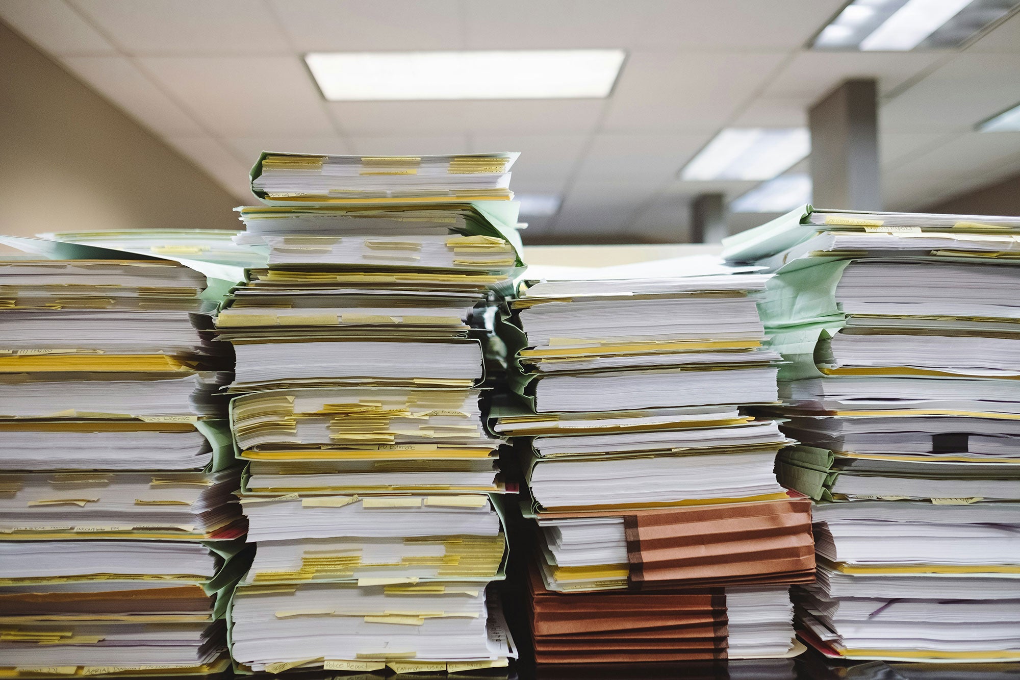 Four tall stacks of papers and file folders under overheard office fluorescent lights.