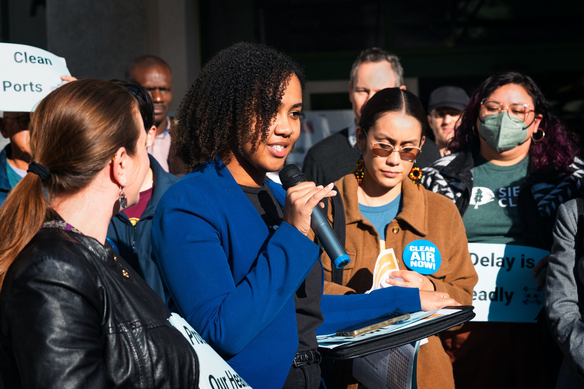 Earthjustice attorney Candice Youngblood, in a bright blue blazer, speaks into a hand-held microphone, surrounded by a large crowd of people on a sunny day.