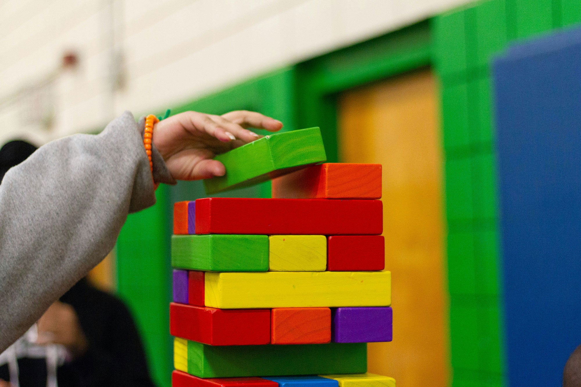 A child's hand places a green building block on top of a tall tower of neatly stacked building blocks.