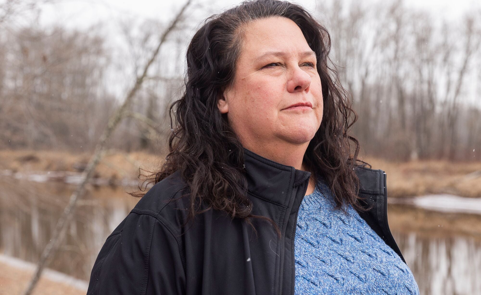 Bad River Band of Lake Superior Chippewa Chairwoman Liz Arbuckle stands next to a river, looking off into the distance. She's wearing a black jacket over a blue knit sweater.