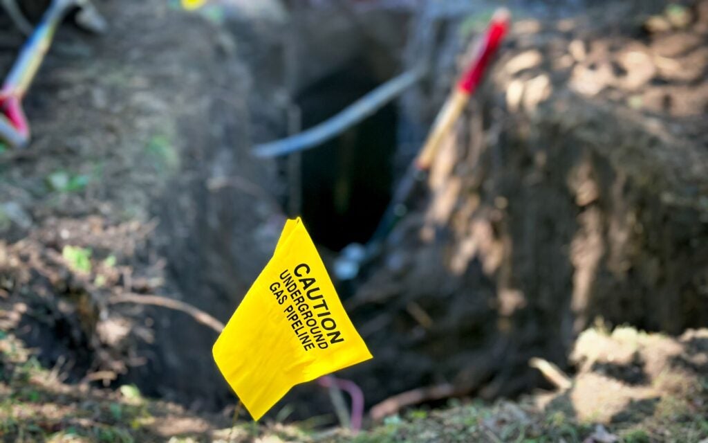 A small yellow flag with the words "Caution: Underground Gas Pipeline" sticks out from a large hole dug into the ground.