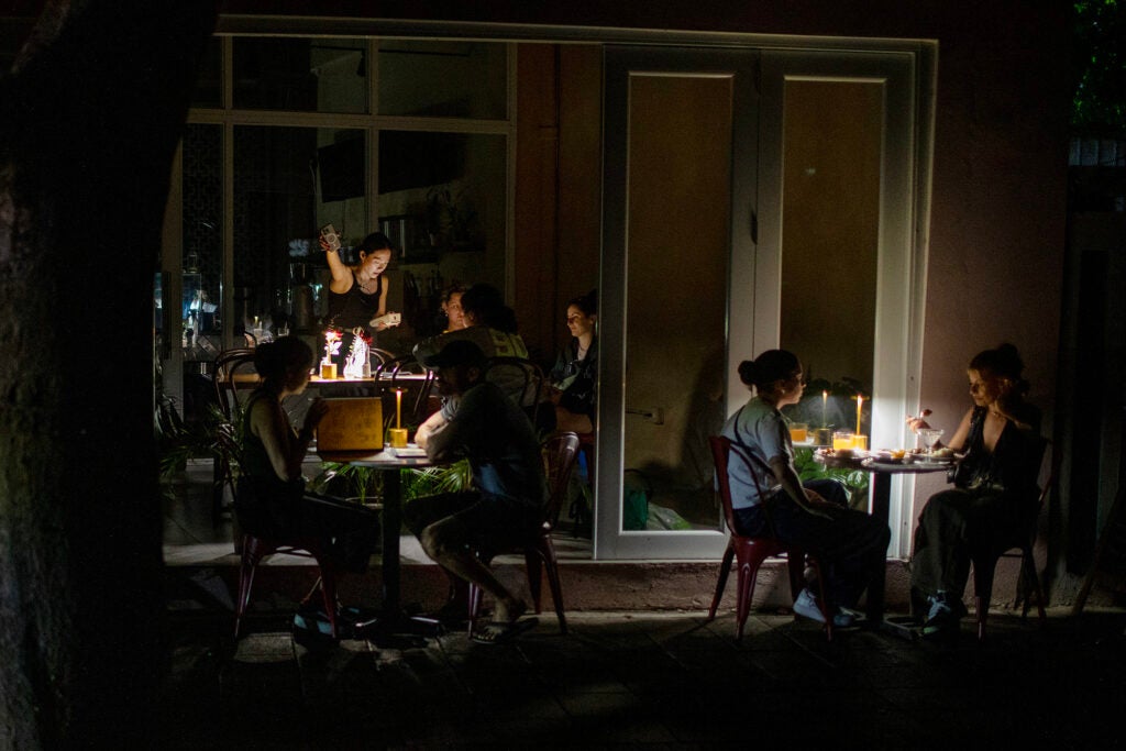 People sit at tables on the sidewalk at a restaurant. The tables are lit by small lamps but the scene is otherwise very dard.