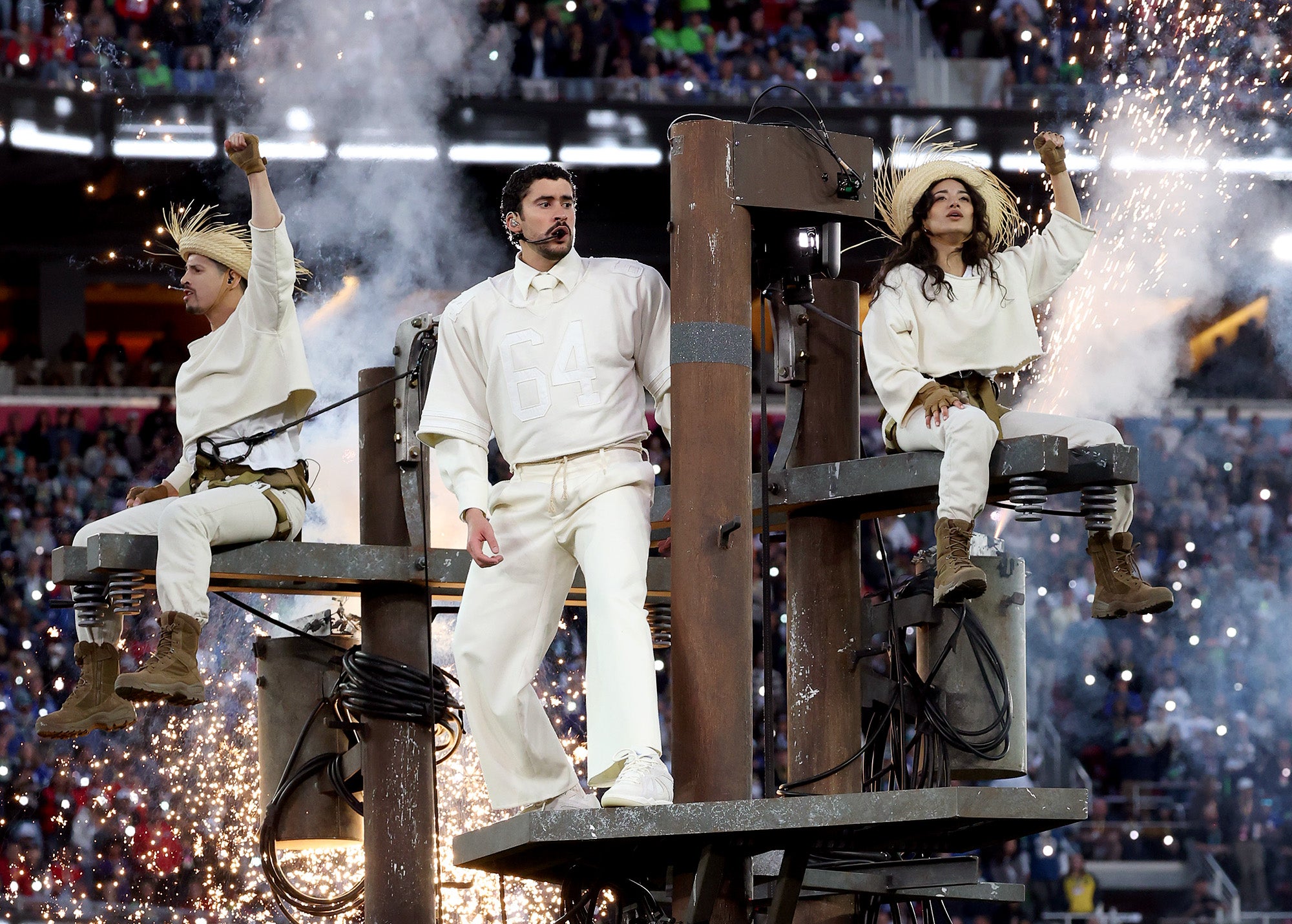 Three people wearing white outfits, the middle one being Bad Bunny, stand and sit on top of utility poles with electrical equipment. The two people on the sides have their fists raised as sparks shoot from below them. The background is a crowd of people in a football stadium.