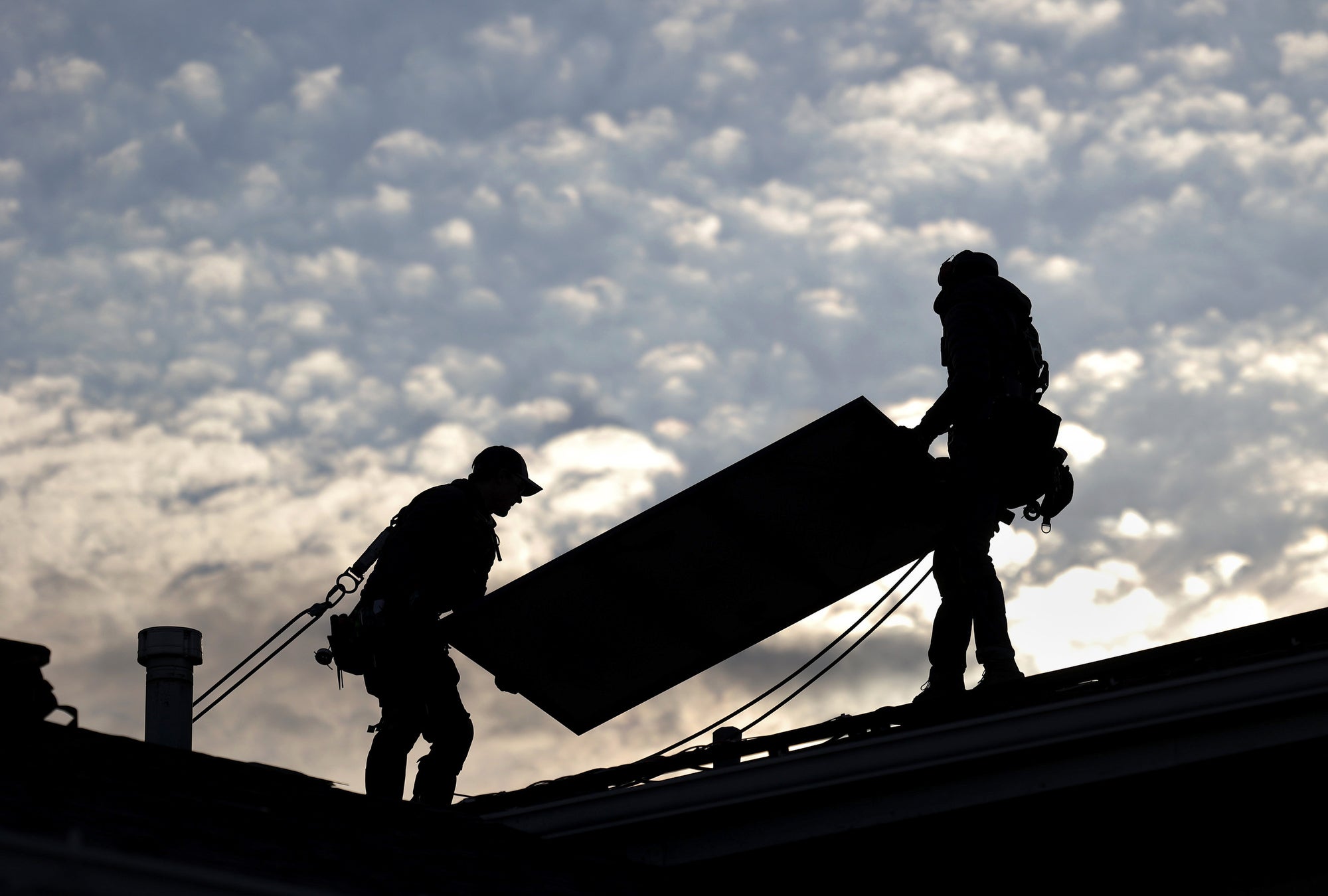 A silhouette of two people carrying a solar panel on a roof.