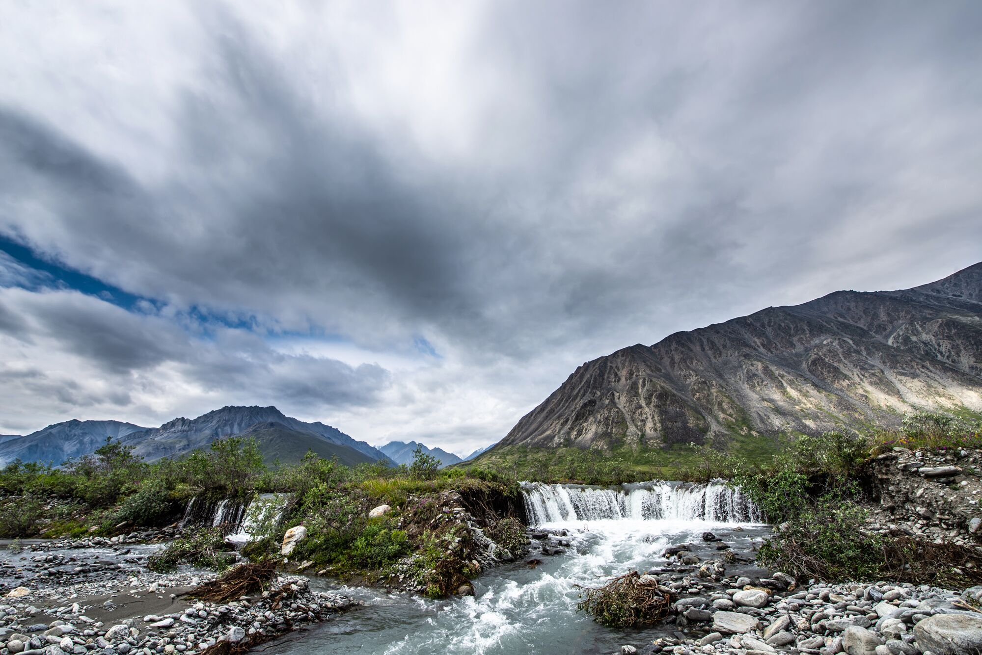 View of waterfall drop-off in Hulahula River and surrounding grassy hills and mountains from the Hulahula River, under an overcast sky.
