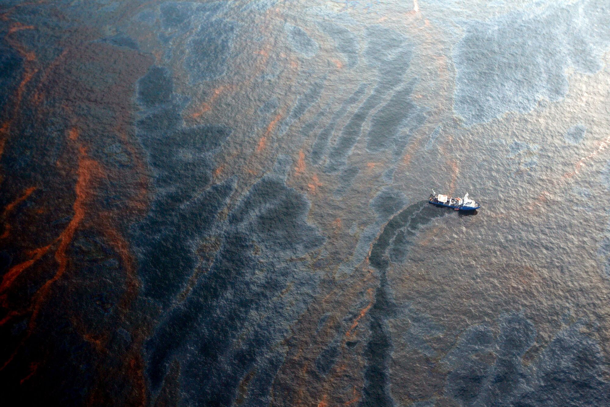 An aerial view of large swathes of brownish-orange oil streaked across the ocean. A small boat is in the middle of the oiled water.