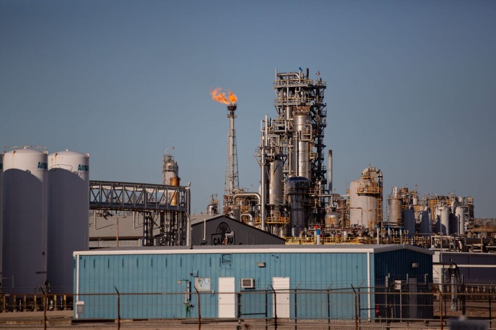 "Dow's Union Carbide Corporation plastics plant in Seadrift, Texas, viewed from outside the facility's perimeter fence. The complex features dense industrial pipework. A gas flare burns near the center of the facility.
