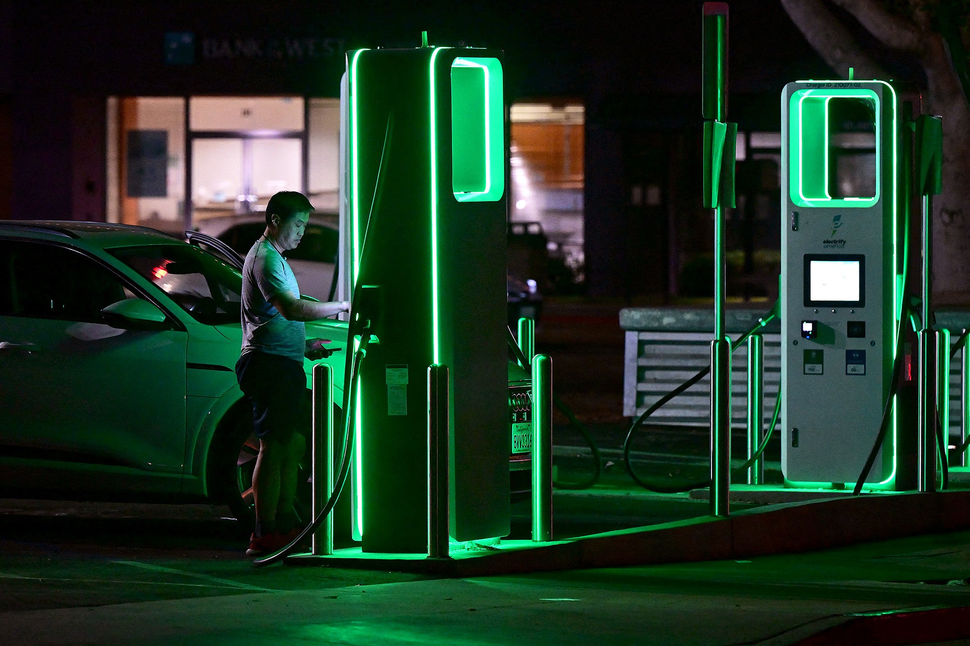 A man interacts with a charging station at night. The stations are covered in green LED lights, illuminating the area.