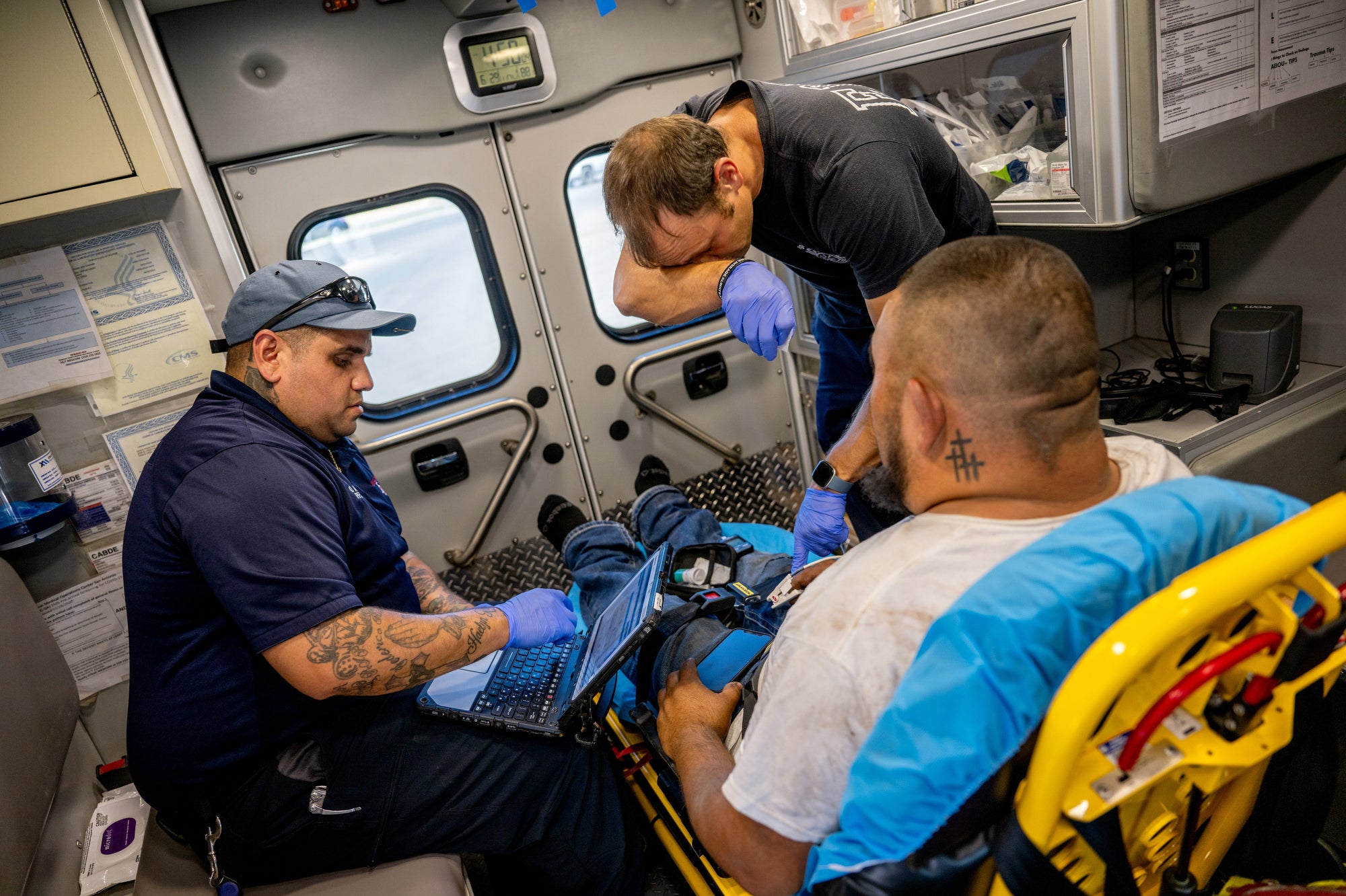Firefighters tend to a man in an ambulance. One is wiping his brow.