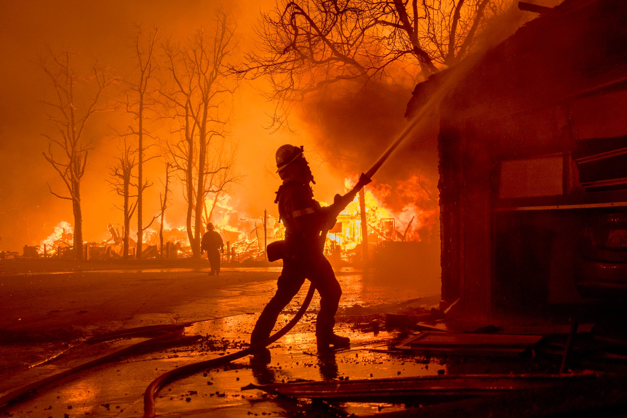 A firefighter sprays a burning home with a hose at night, surrounded by orange burning homes.