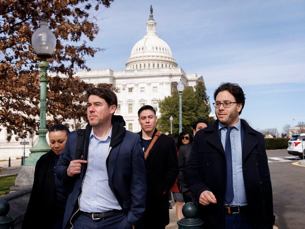 A group of people walk along a sidewalk with the dome of the US capitol visible in the background.