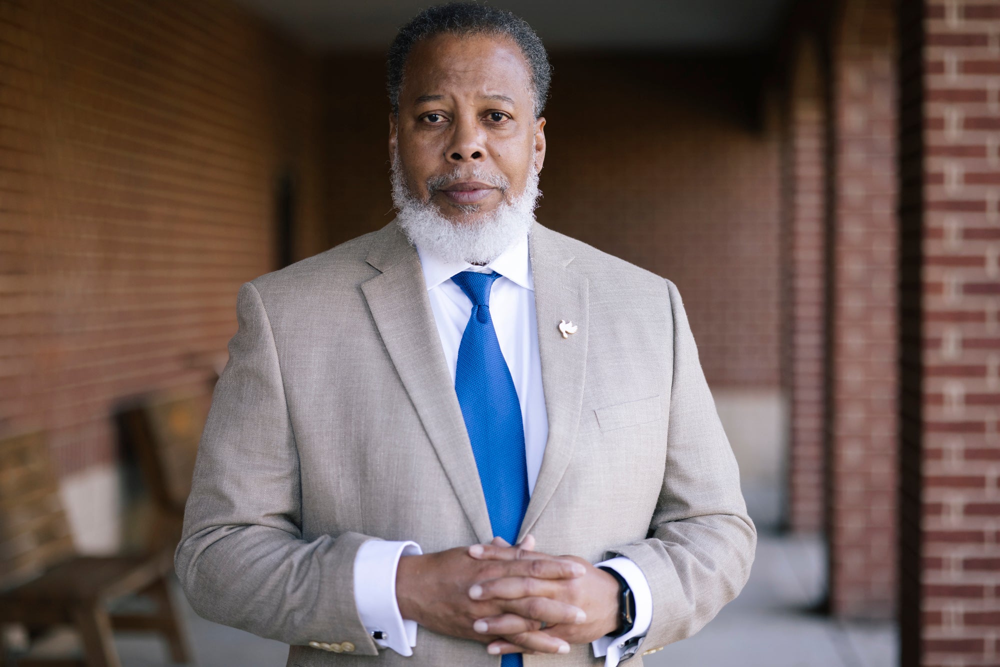 A man in a light gray suit and blue tie looks directly into the camera with his hands clasped.