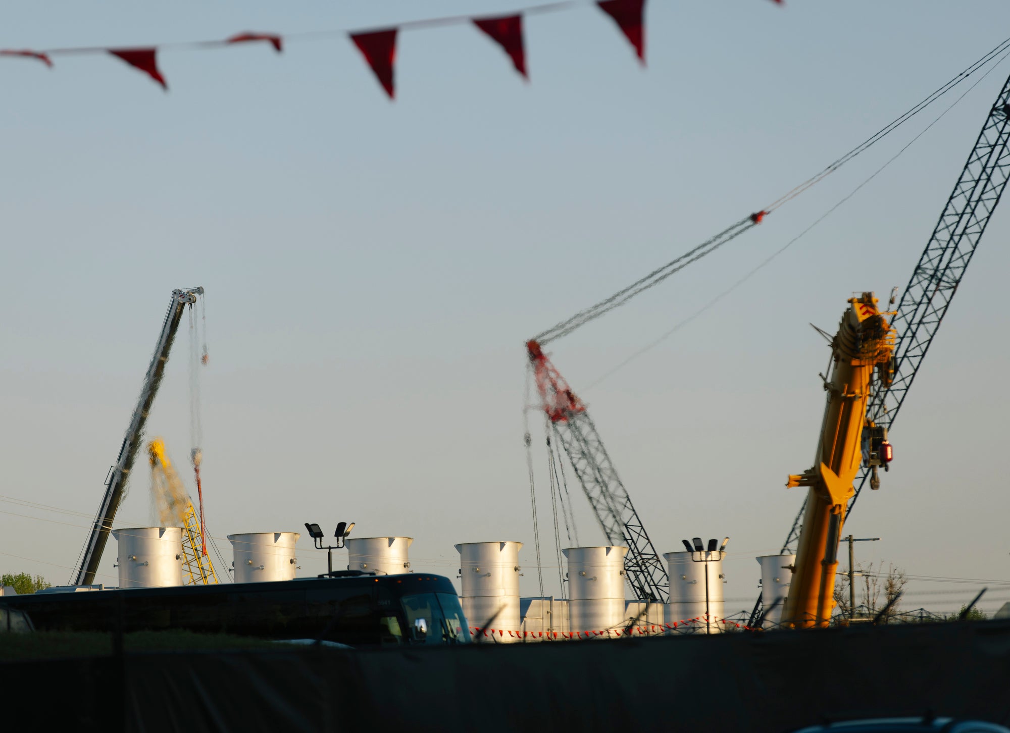 Seven large, silver exhaust stacks are visible over a fence. They are operating with visible heat coming out the top. They are also surrounded by construction cranes.