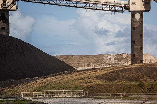 Industrial coal ash site with large gray mounds of waste, a conveyor structure overhead, and barren, brown terrain under a cloudy sky.