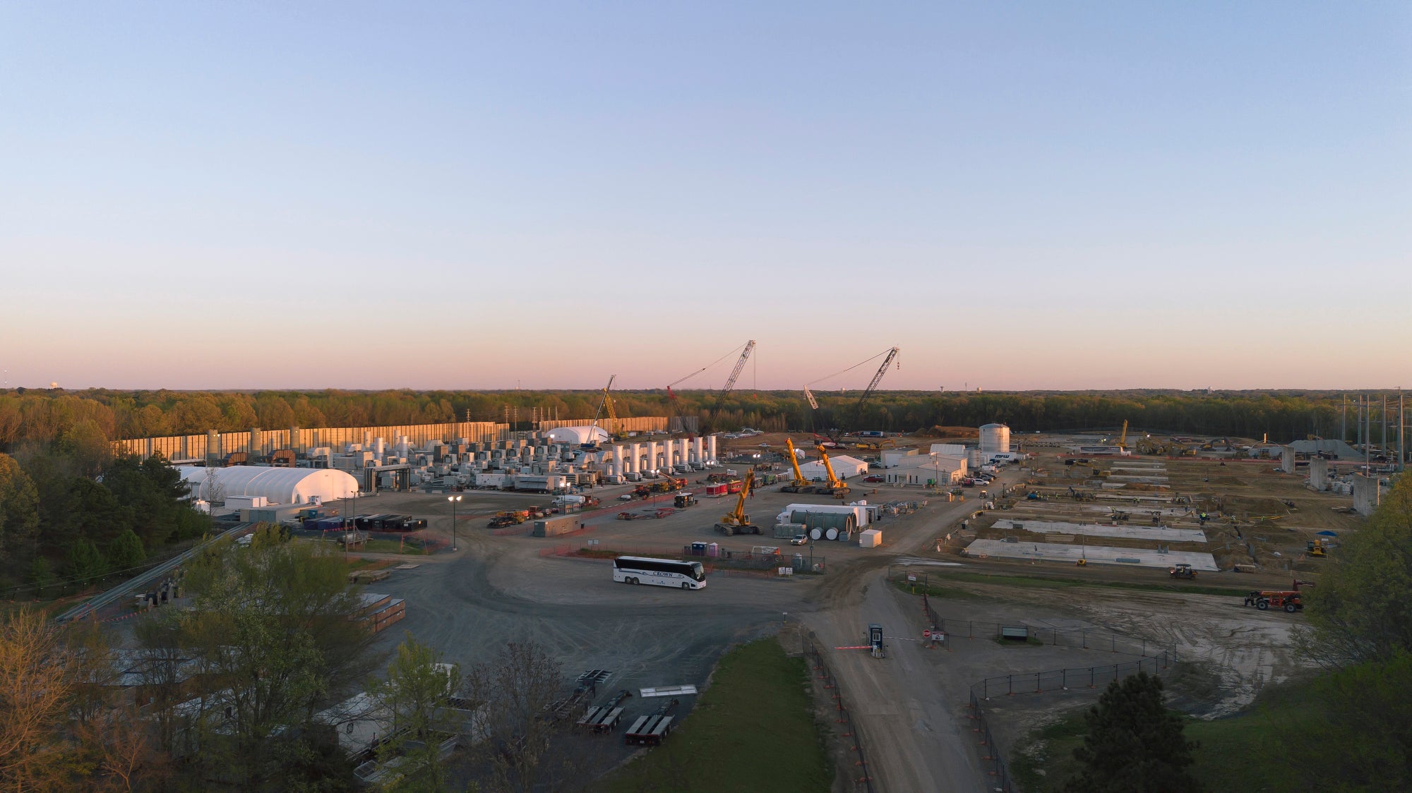 A wide aerial landscape photo of a construction site with gas turbines operating on the left side of the area. The site is surrounded by trees in the distance.