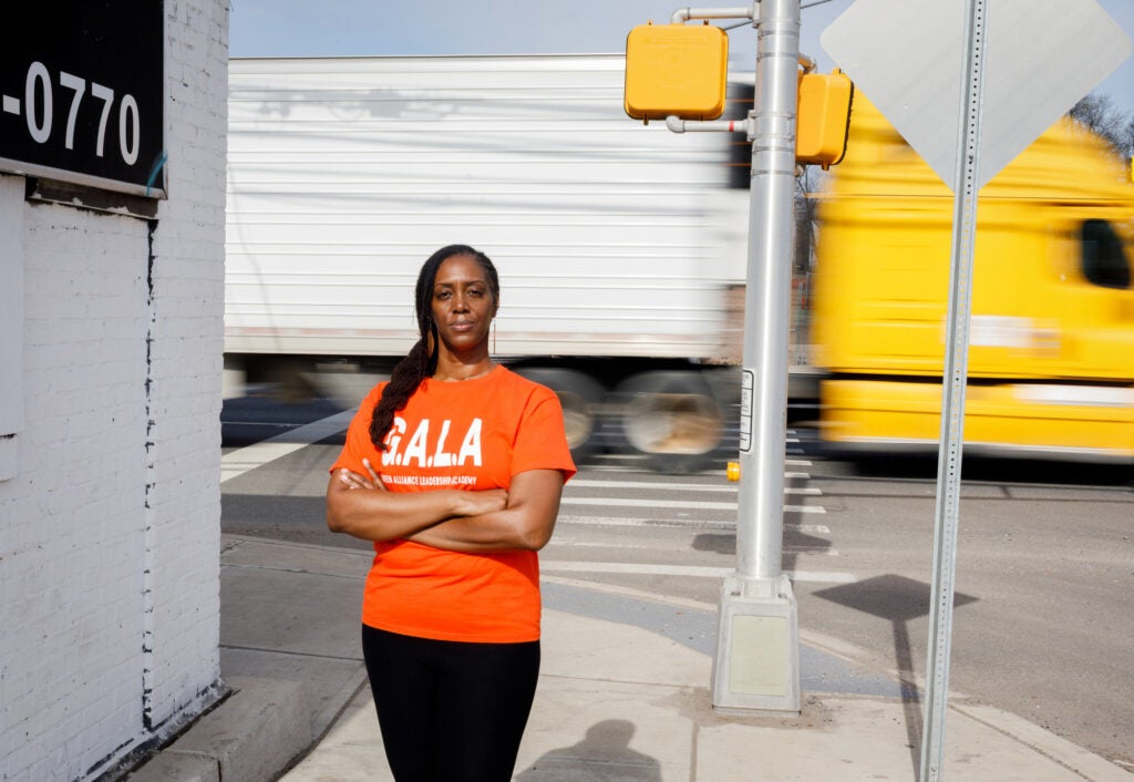 A woman in an orange t-shirt stands with her arms crossed looking directly at the camera as a blurry semi truck passes behind her.