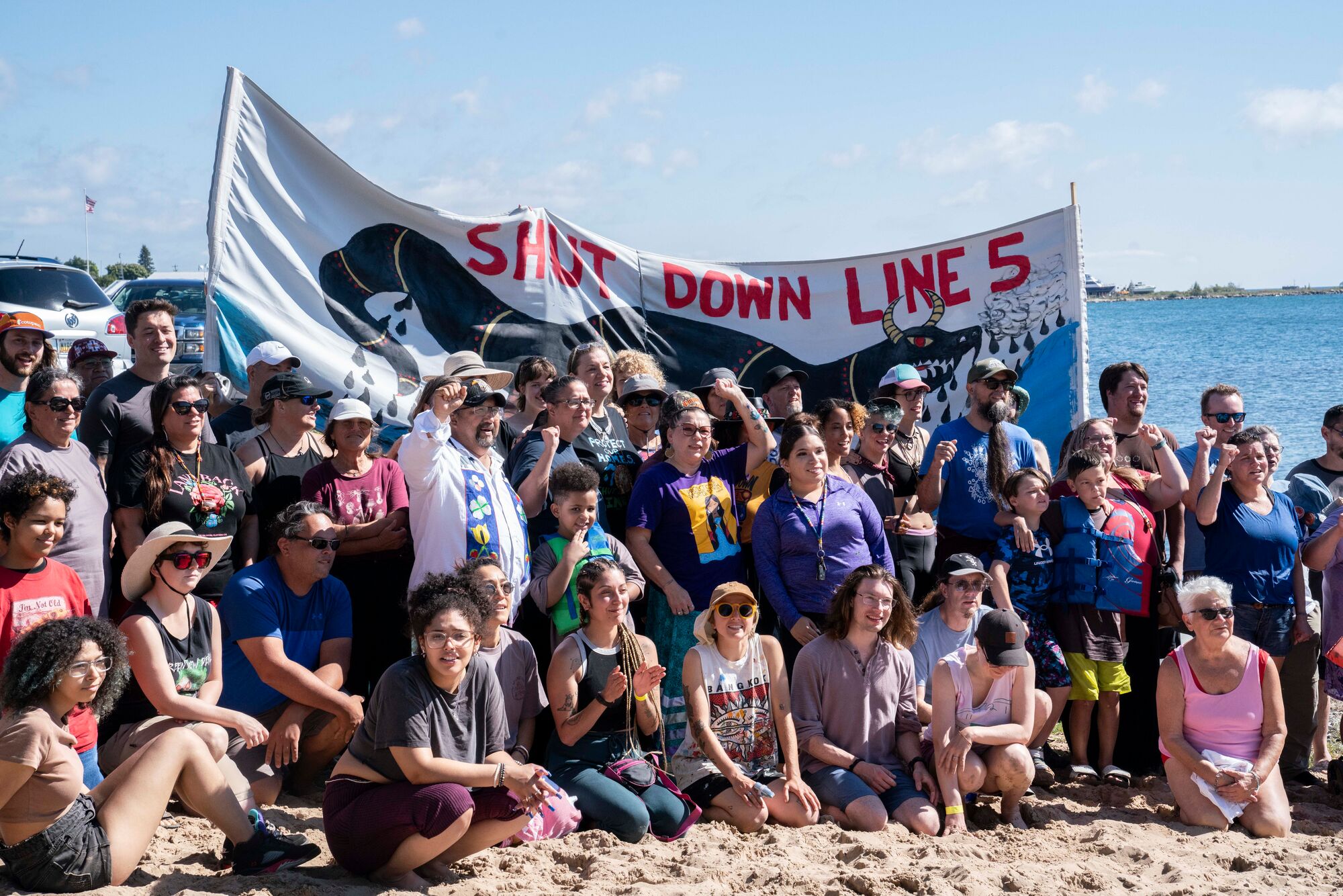 A large crowd of people pose on the shores of a lake on a warm, sunny day. A large banner is setup behind them that reads, "Shut Down Line 5."