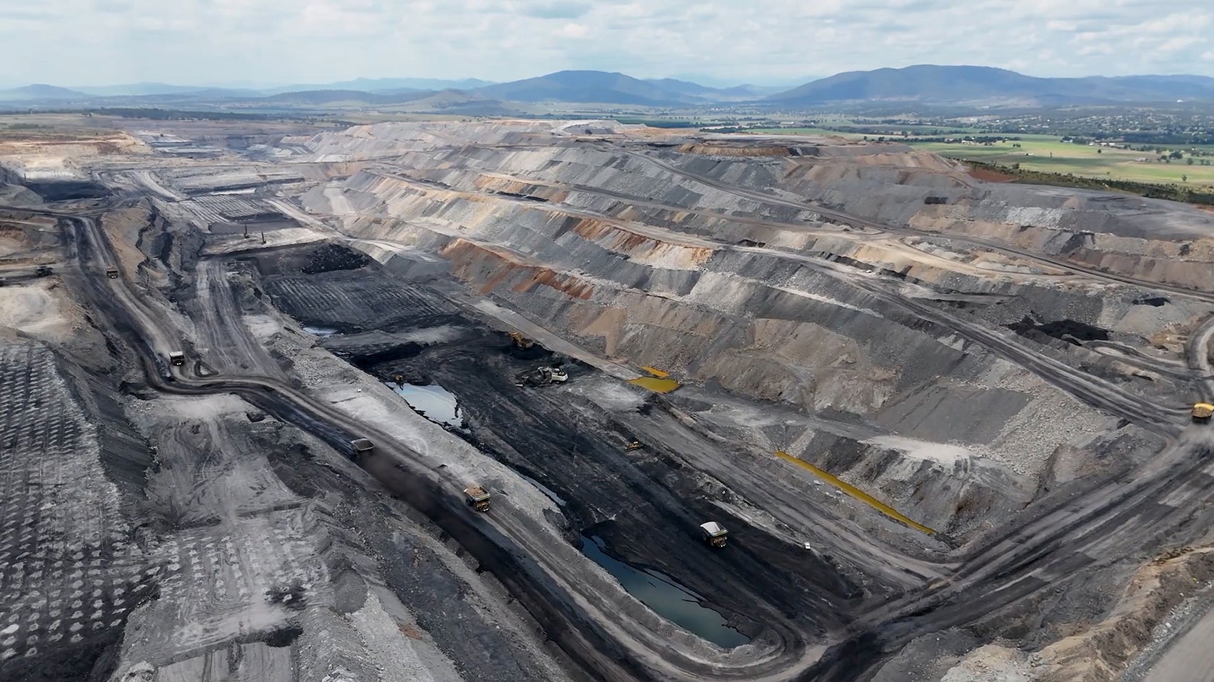 A large, gray, open pit mine surrounded by green fields and low mountains.