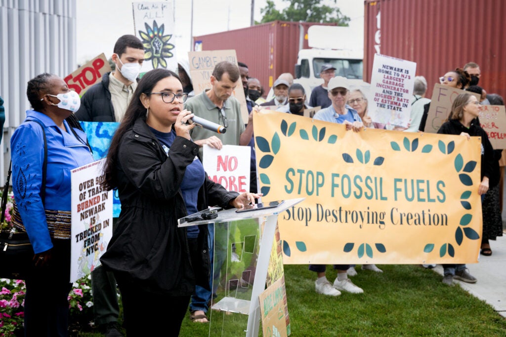 A woman holding a microphone and standing at a small podium talks as a crowd of people stand behind her. Some are holding signs. A large one reads "Stop fossil Fuels. Stop destroying creation".