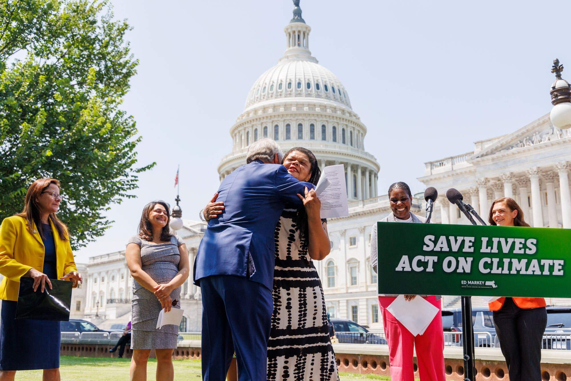 Rep. Paul Tonko (D-NY) embraces Leslie Fields of WE ACT for Environmental Justice near a podium with the sign, "Save Lives. Act on Climate." They are flanked by two people on either side. The group is standing in front of Capitol Hill on a bright, sunny day.