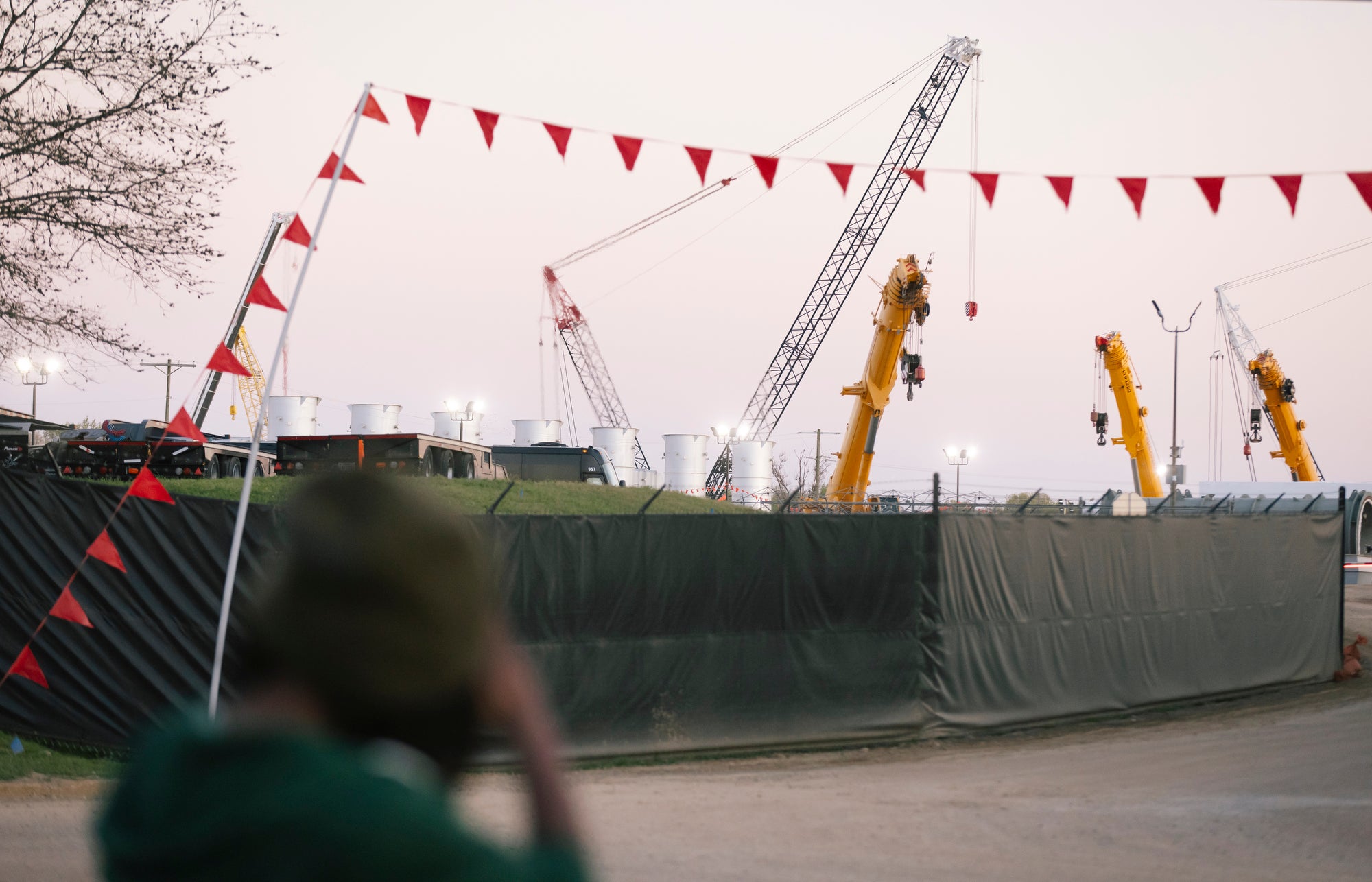 A blurry figure in the foreground looks at the construction of gas turbines behind a tarp-covered fance.