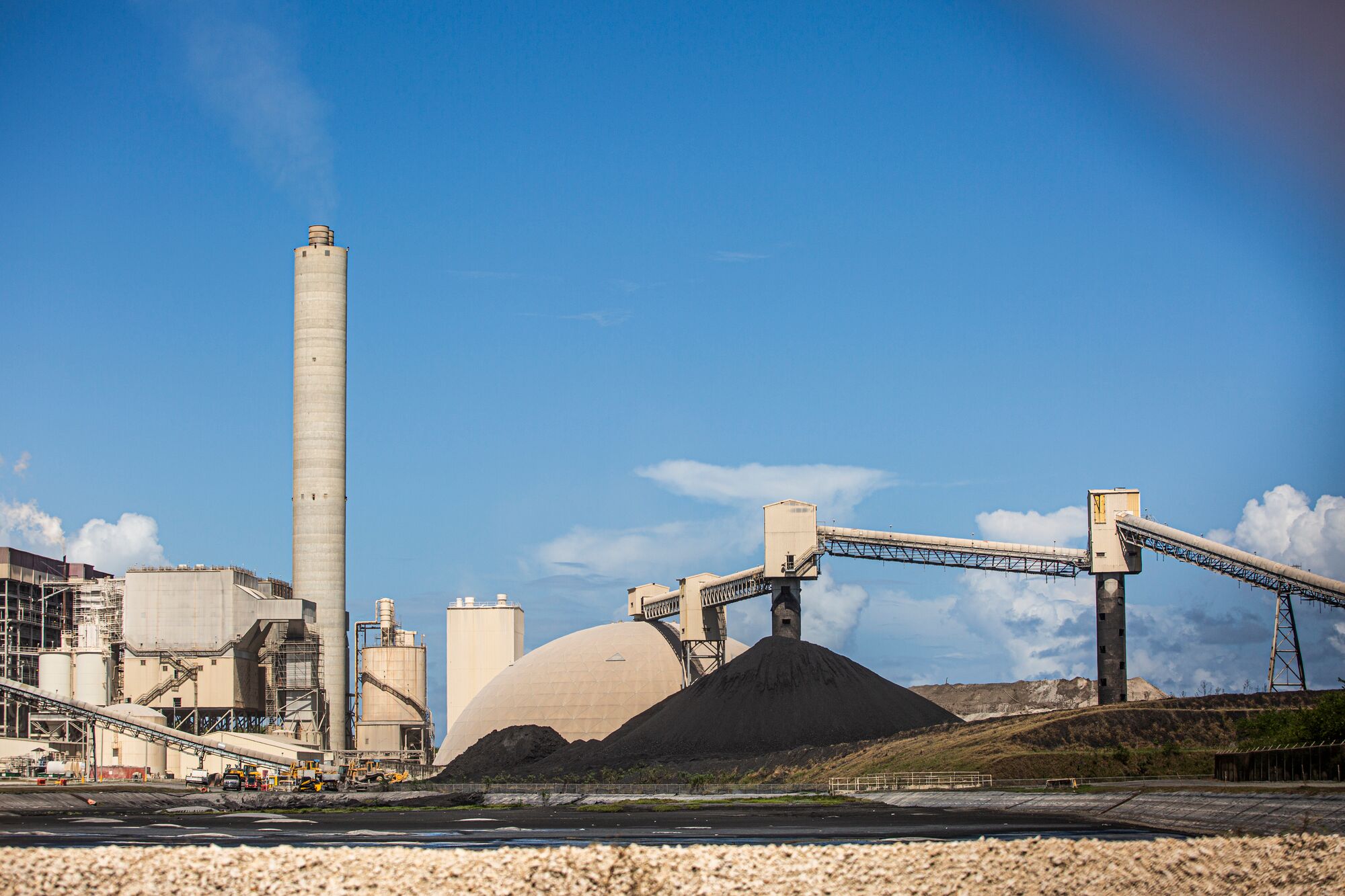 A sprawling industrial facility with a large, uncovered mountain of black coal in the mid-ground and a mountain of light grey toxic coal ash behind it.