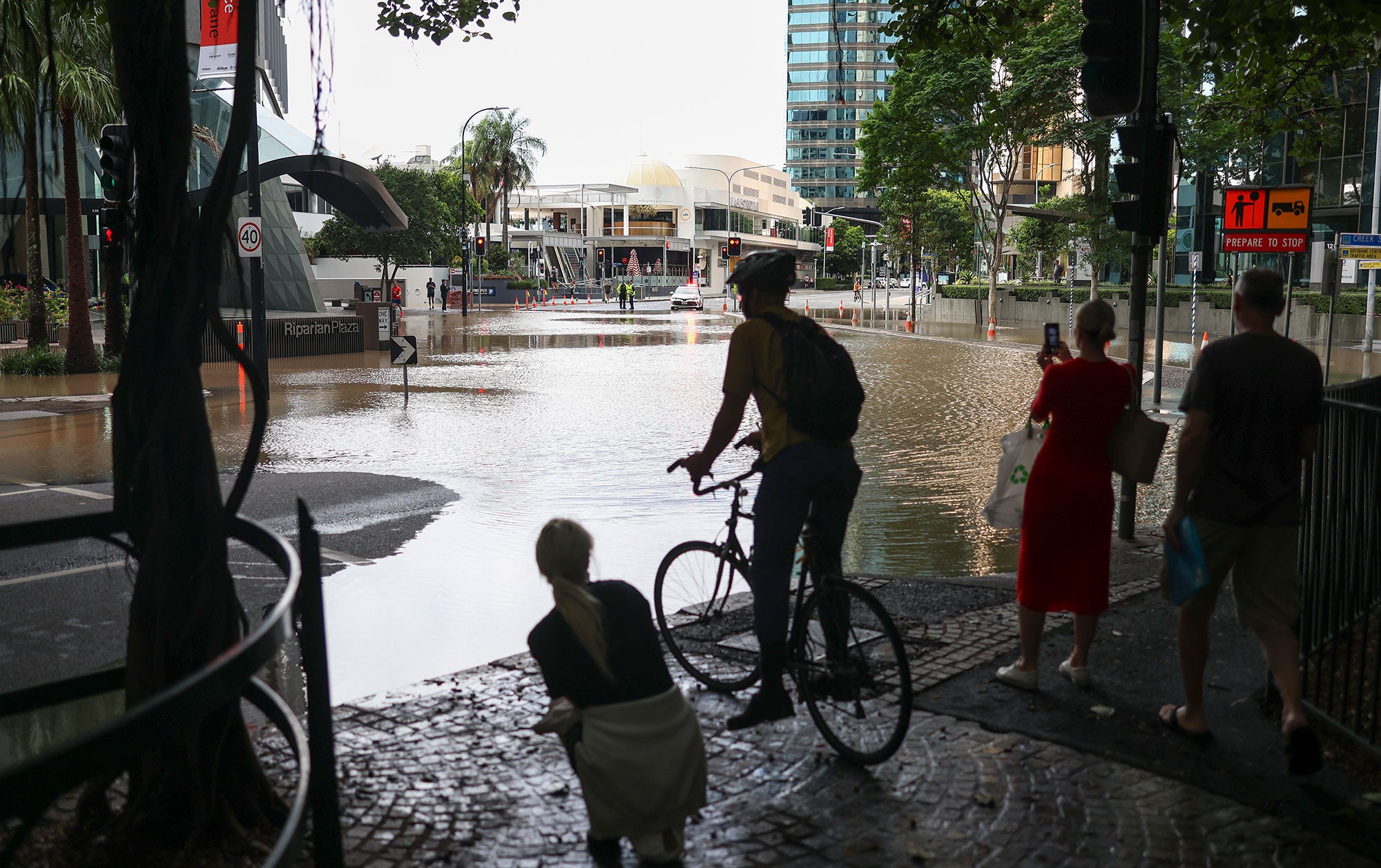 Three pedestrians and one bicyclist are standing at the edge of floodwaters, with their backs to the viewer, looking out onto a completely submerged roadway in a business district. One of the pedestrians, wearing a red dress and holding a plastic shopping bag, is using her mobile phone to capture the scene. Two law enforcement people are visible in the far distance.