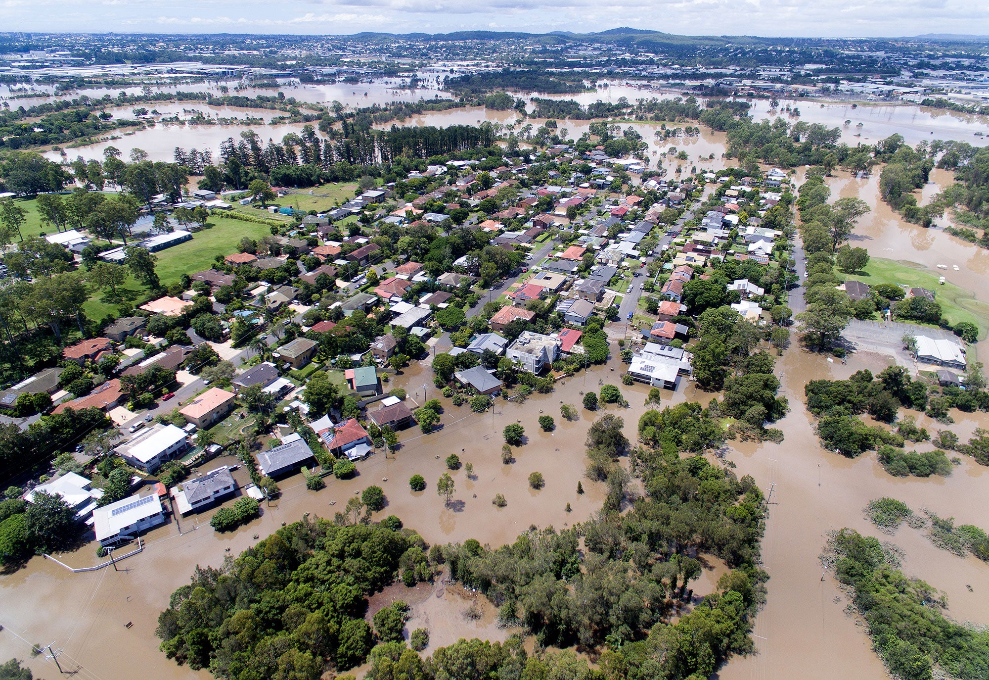 Aerial view of dozens, perhaps hundreds, of homes, partially submerged in murky, brown water. The floodwaters seem to stretch for miles. Bright green trees are visible above the floodwaters.