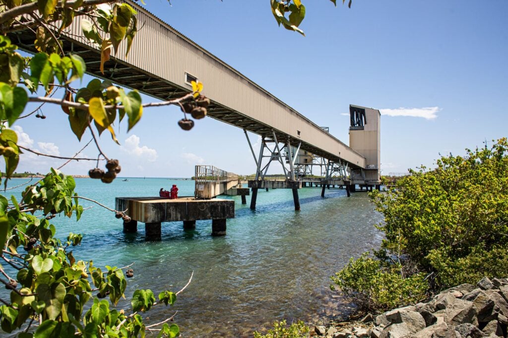 A large industrial structure sits in clear, light blue waters along a rocky coast. An elevated tunnel stretches from the structure's outpost to the land.