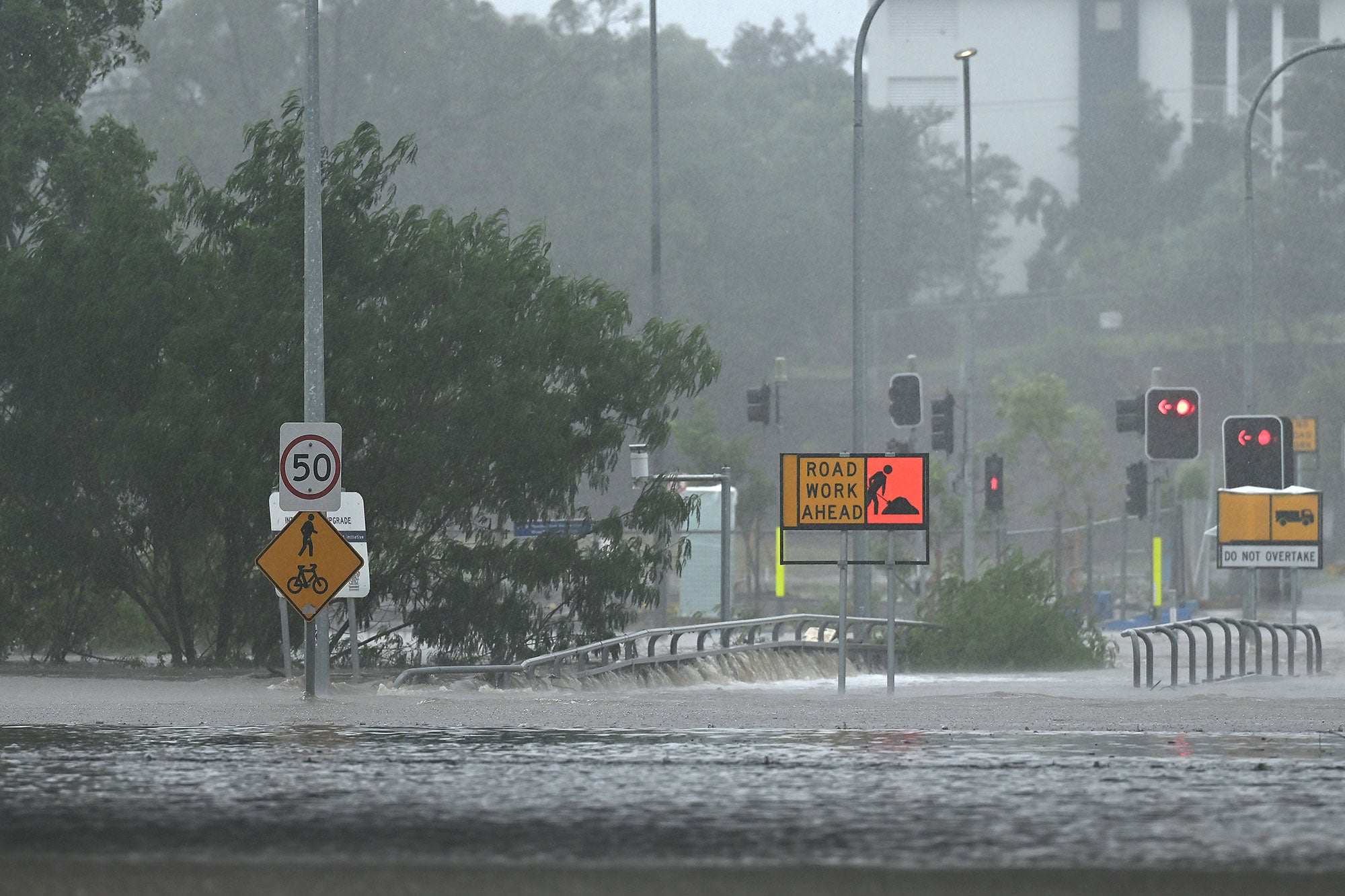 Torrential rain is coming down on an urban road, adding to the floodwaters that are already rising halfway up the road sign posts and traffic lights. The traffic lights are all red.