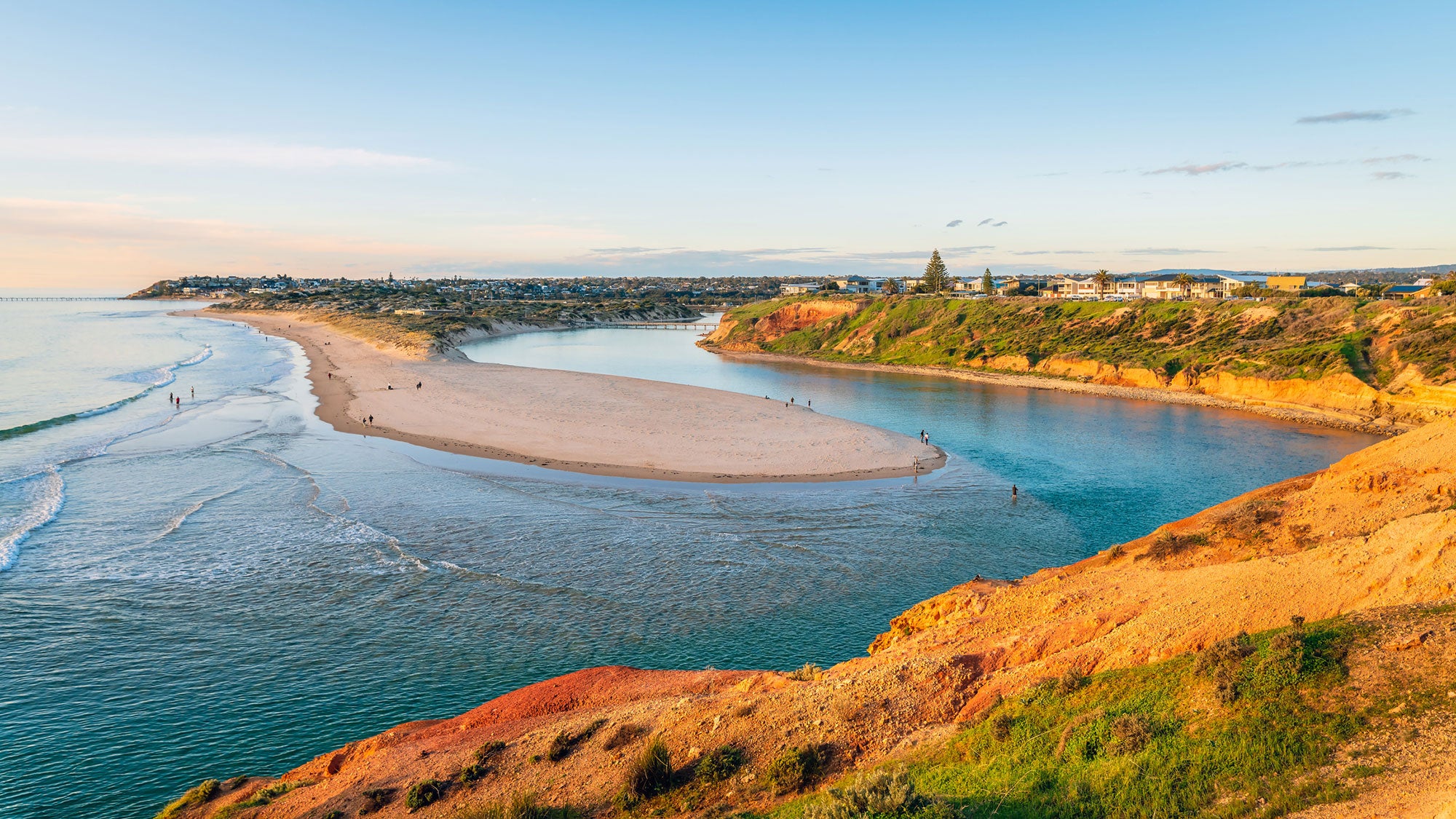 An elevated view of the blue-green waters of the ocean, with gently lapping waves on the left. A river mouth curves around a sandy promontory from the right to meet with the ocean waters. Several people, looking very tiny, are on the beach and in the waters. In the hear foreground is a hill with vegetation bathed in warm, golden light. In the distance on the right and in front are densely packed homes. The sky is a light blue with a few wisps of grey clouds.