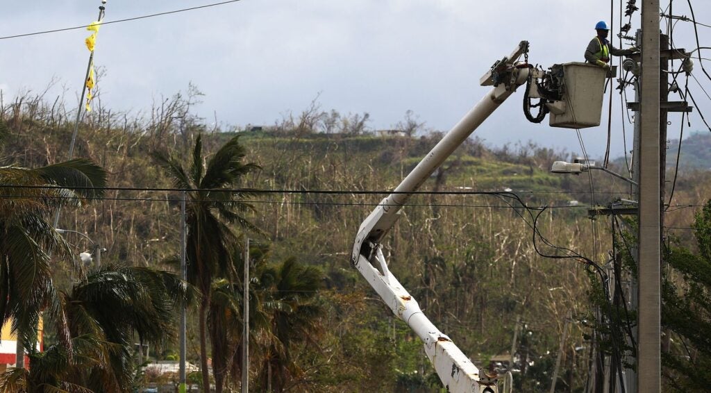 A worker in a yellow safety vest is inspecting the top of a power line pole from the bucket of a cherry picker truck, with its arm almost fully extended. In the background, a once-lush landscape is severely denuded of vegetation.