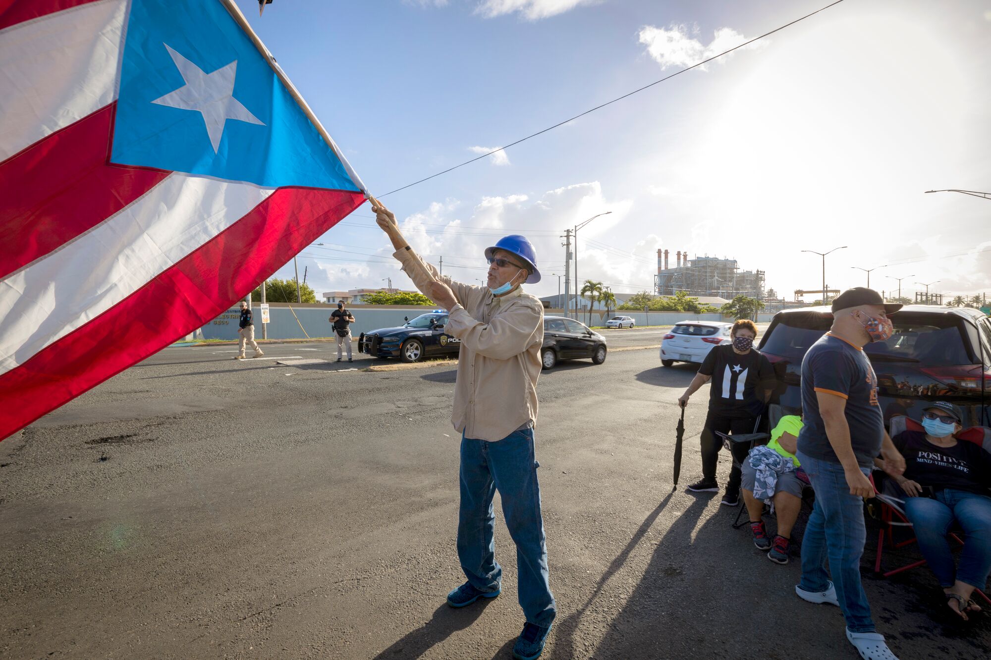 A man in jeans, beige button-up shirt, and bright blue hard hat vigorously waves a huge Puerto Rico flag on the side of a street under a bright, sunny sky. A number of other people (some of them wearing protective face masks are standing and sitting next to vehicles near the man. Cars are driving through the street. A sizeable, heavy industrial facility is clearly visible on the other side of the street.