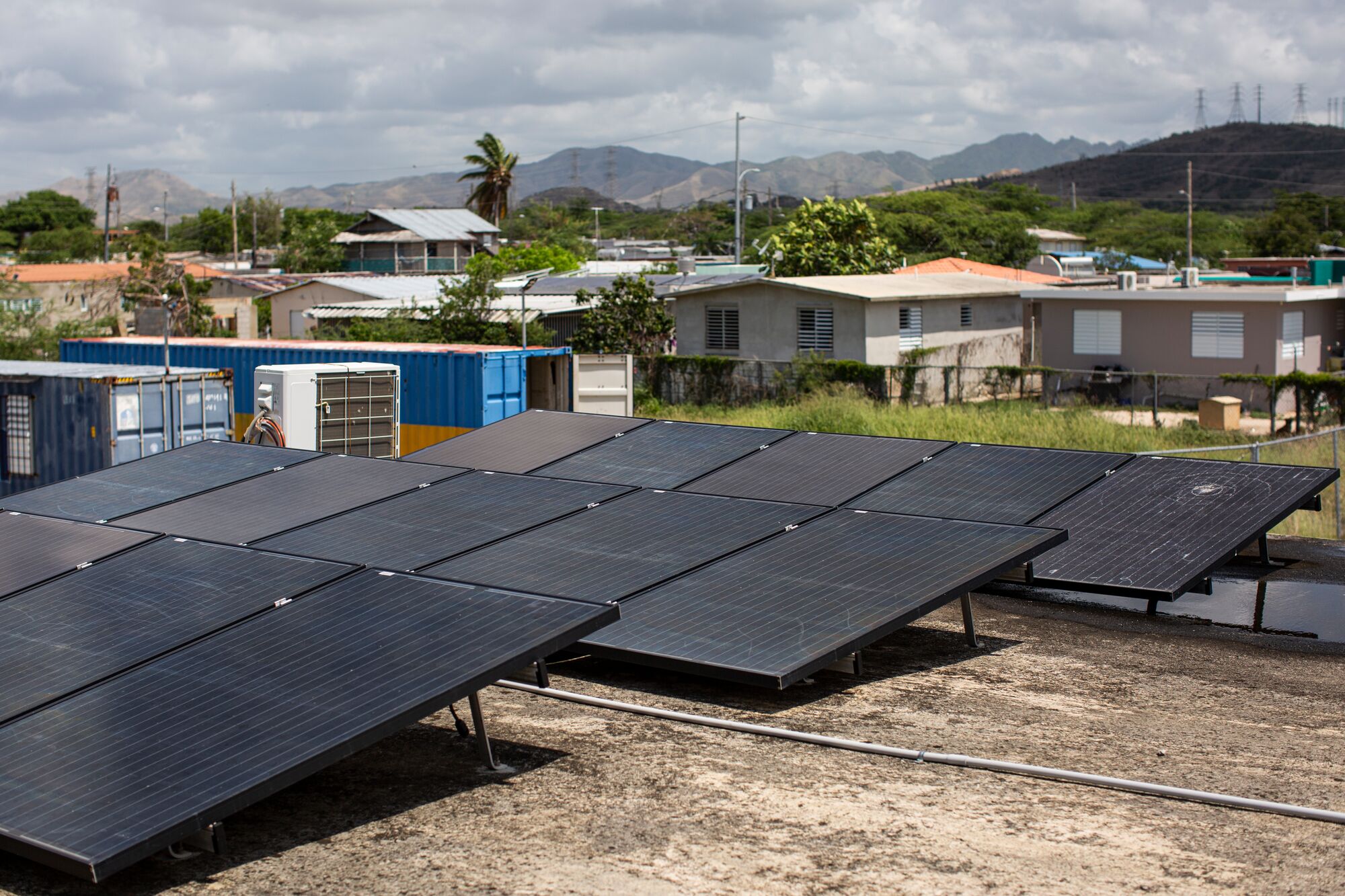 Three rows of several solar panels on a flat roof, with shipping containers and homes on the ground-level. Transmission towers are visible in the distance along the tops of hills.