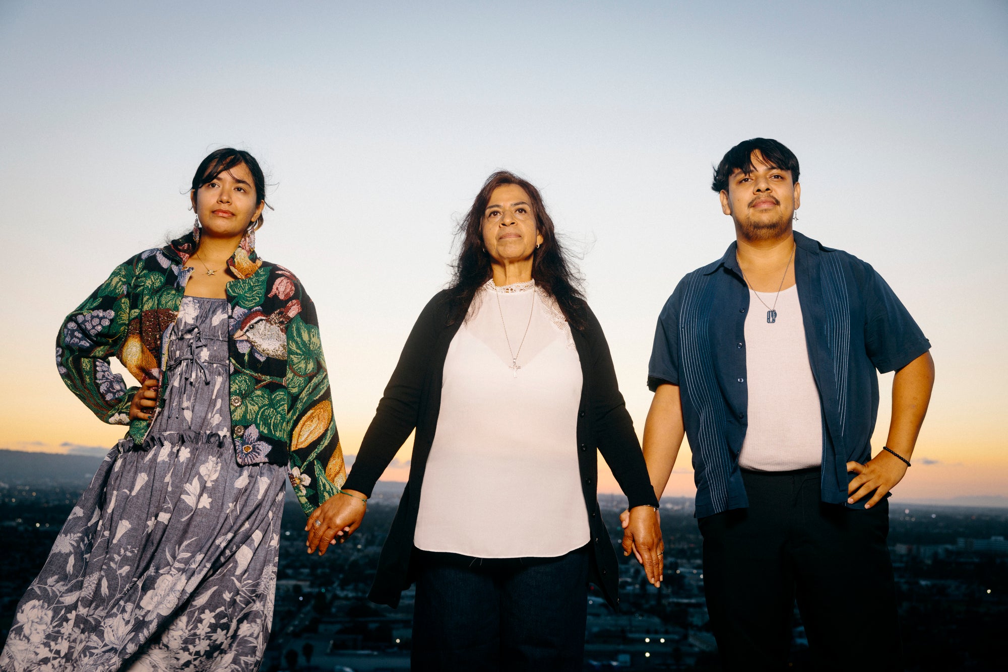 Three people hold hands and stand for a portrait while looking into the distance, towards the camera. A city landscape can be seen behind them.